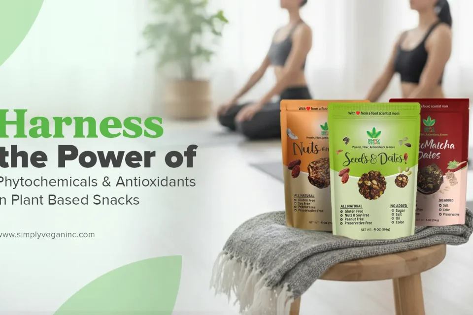Three Simply Vegan snack packs of Nuts on Dates, Seeds and Dates, and Chocomatcha Dates, displayed on a stool in a bright yoga studio, with two people practicing yoga in the background.
